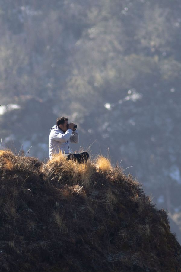 Sherab Dorji, a senior birding guide for Wild Nature Quest, sitting on a grassy Himalayan ridge using binoculars to scan for birds during a tour in Bhutan.